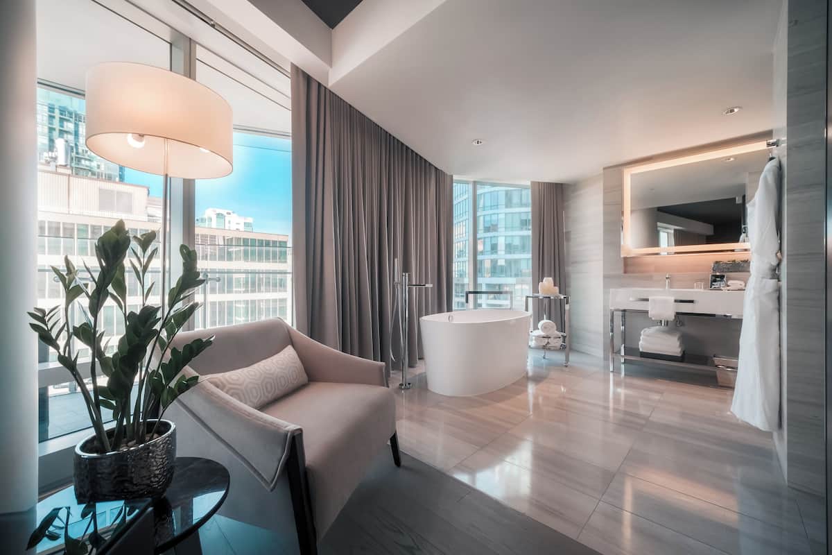 Una habitación de hotel moderna con un ventanal grande, una zona de estar con sofá, una bañera redonda y un tocador con espejo.