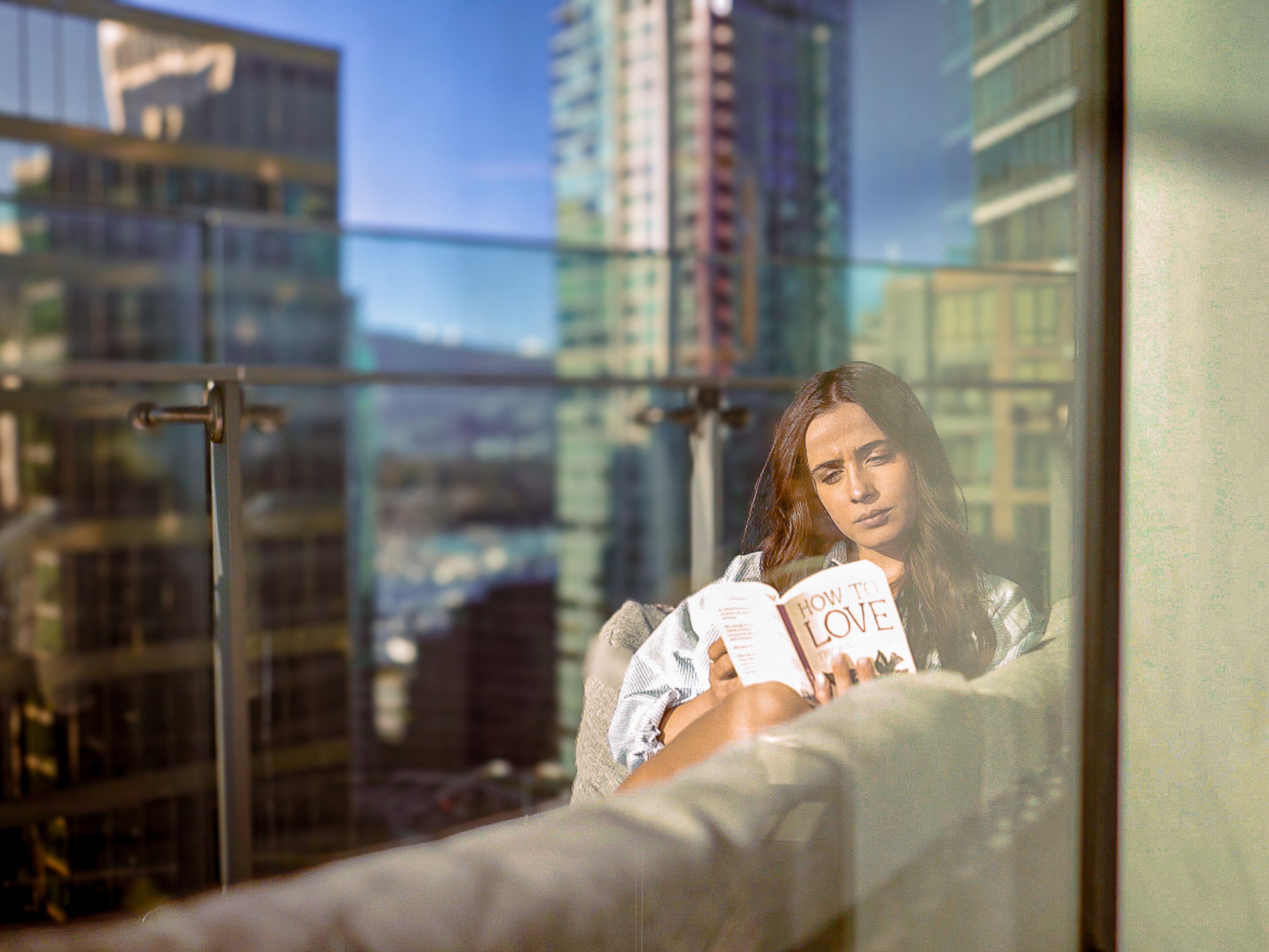Una persona leyendo un libro titulado "Cómo Amar" en un balcón con vistas a la ciudad.