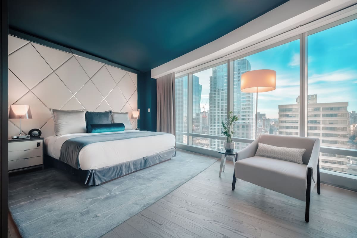 Un dormitorio moderno con una cama grande, mesitas de noche, una silla y vistas de la ciudad a través de amplios ventanales.