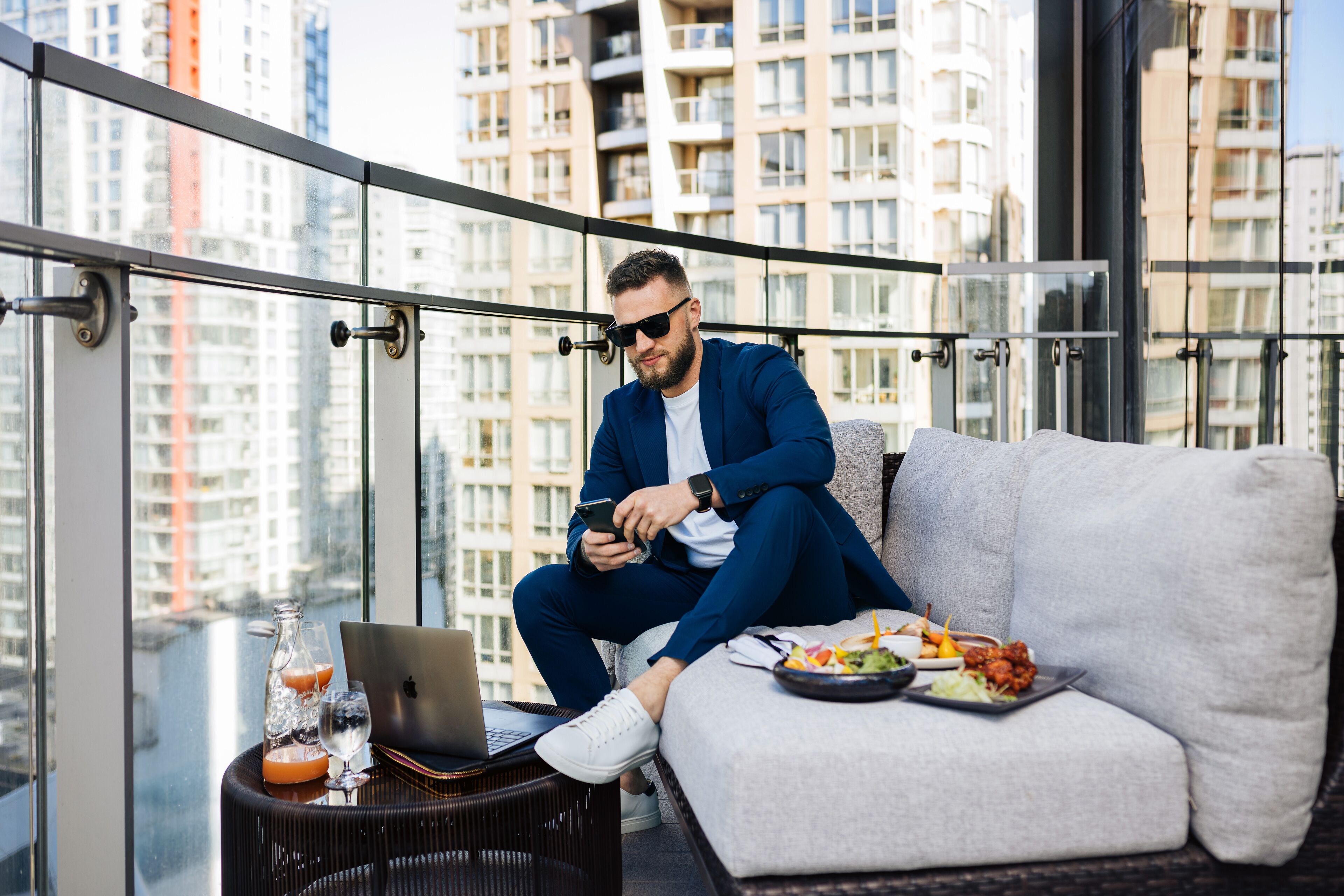 A man sitting on a balcony with a laptop, smartphone, and food on a table.