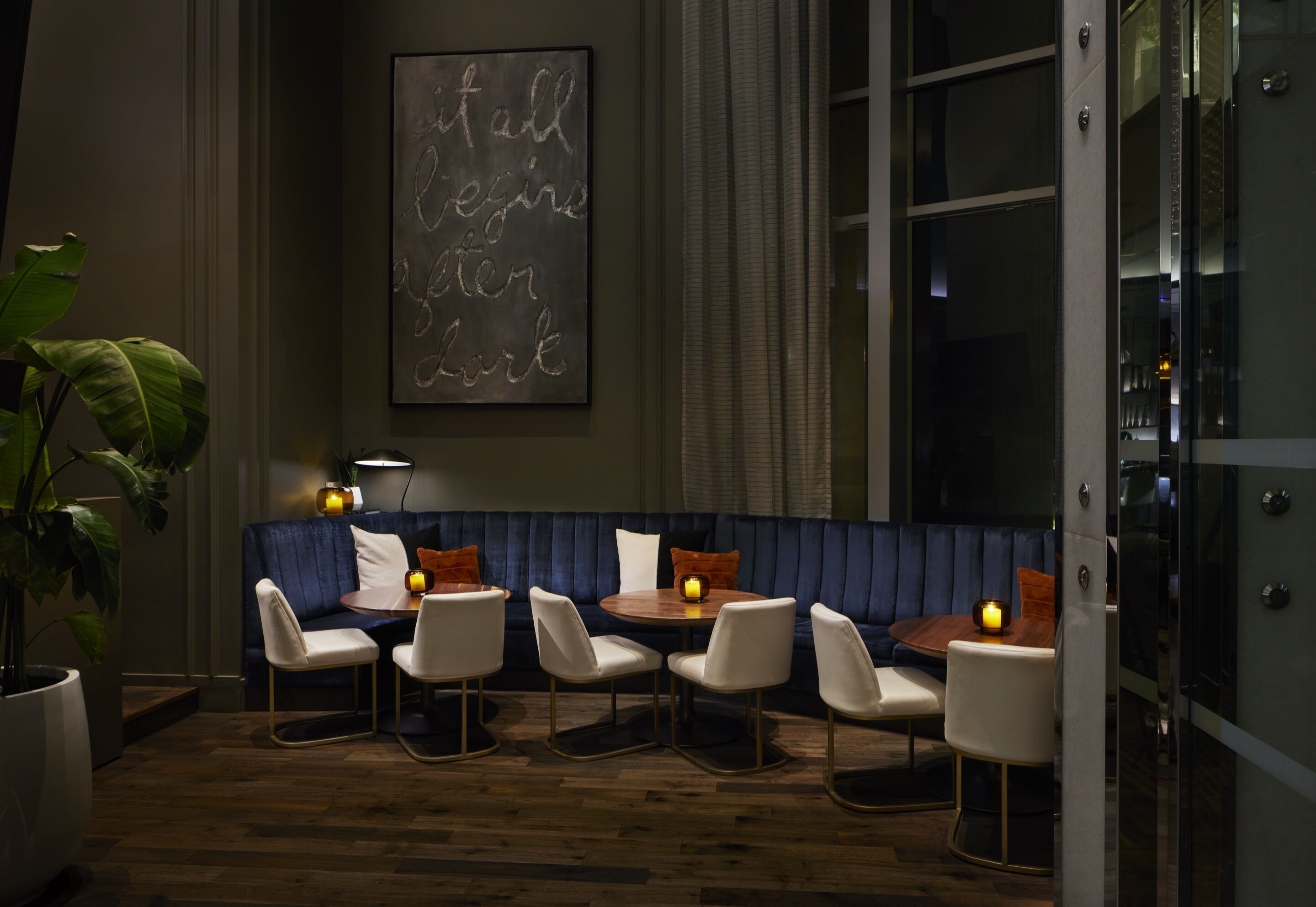 Un coin salon faiblement éclairé, avec un banc bleu rembourré, des chaises blanches et une petite table ronde sur laquelle sont posées des bougies. Un grand panneau encadré est accroché au mur, et une plante est visible dans un coin.