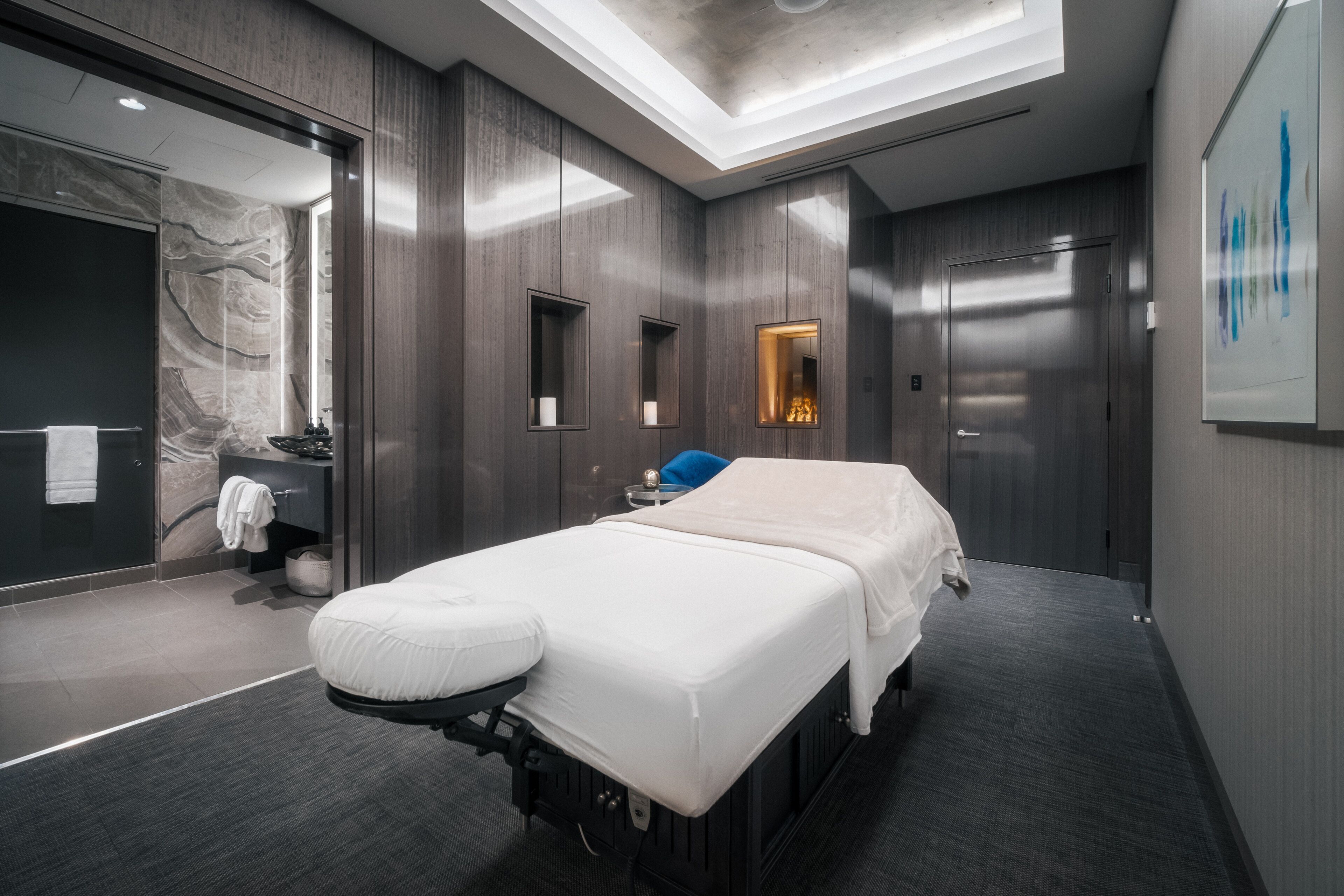 Una habitación moderna y bien iluminada con cama, mesa de masajes, baño pequeño y espejo grande.