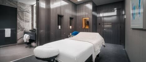 A modern, well-lit room with a bed, a massage table, a small bathroom, and a large mirror.