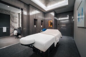 Una habitación moderna y bien iluminada con cama, mesa de masajes, baño pequeño y espejo grande.