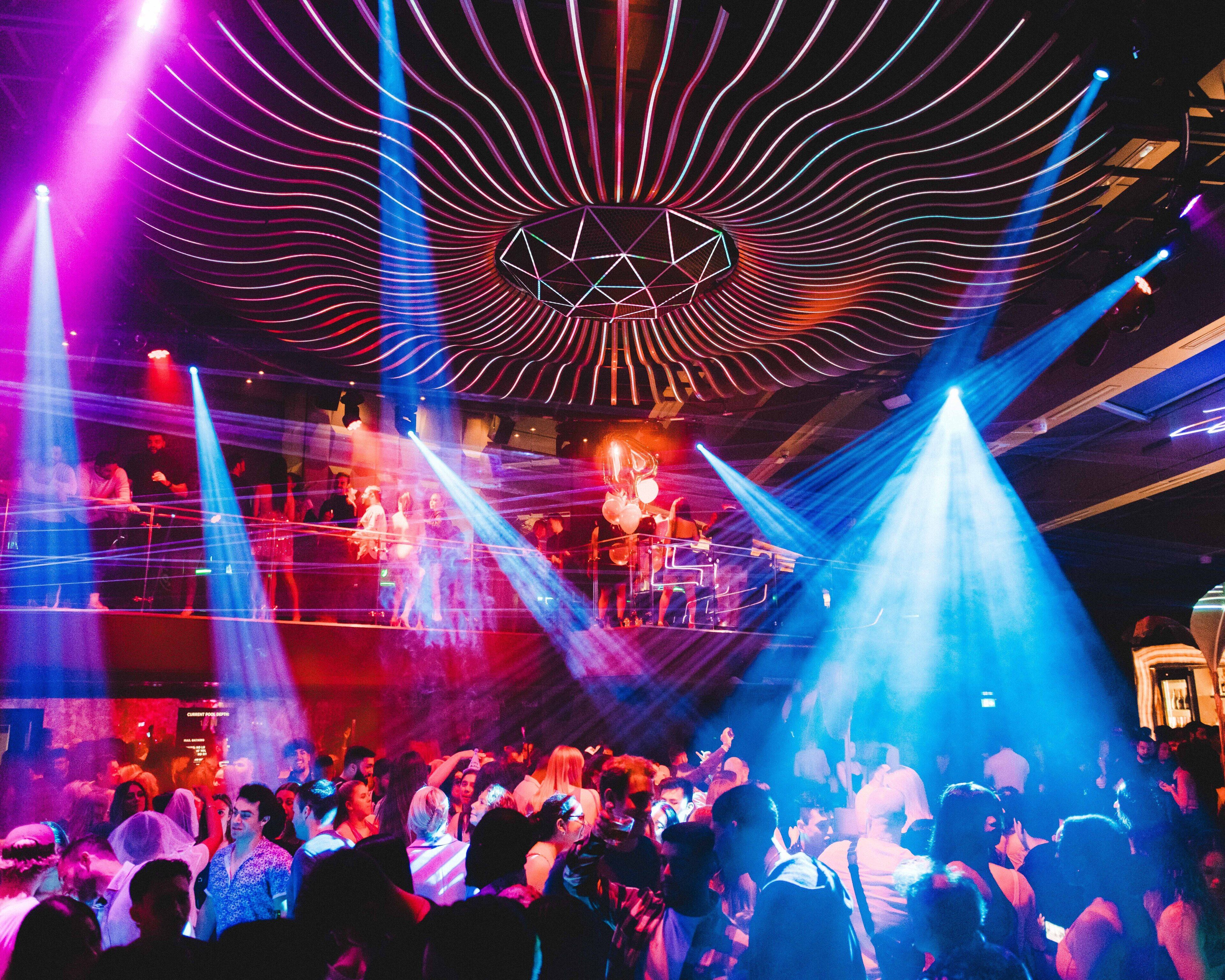 A crowded nightclub with a stage and DJ booth, featuring a geometric light design on the ceiling.