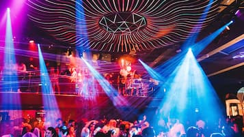 A crowded nightclub with a stage and DJ booth, featuring a geometric light design on the ceiling.
