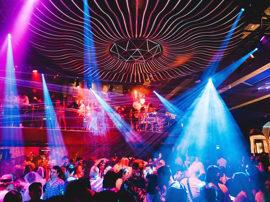 A crowded nightclub with a stage and DJ booth, featuring a geometric light design on the ceiling.