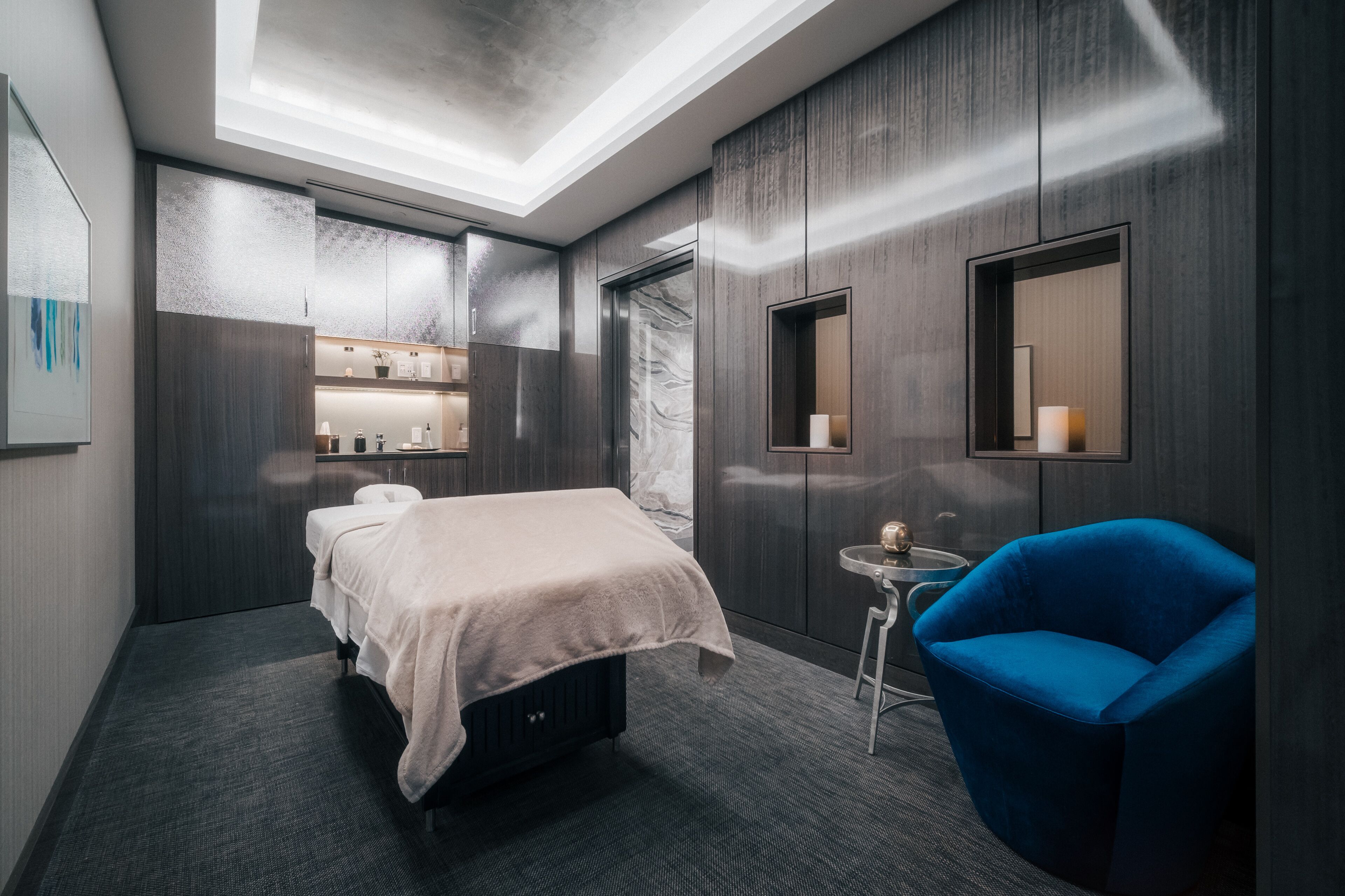 Une chambre d’hôtel moderne avec un lit, un fauteuil bleu, une petite table et un miroir.