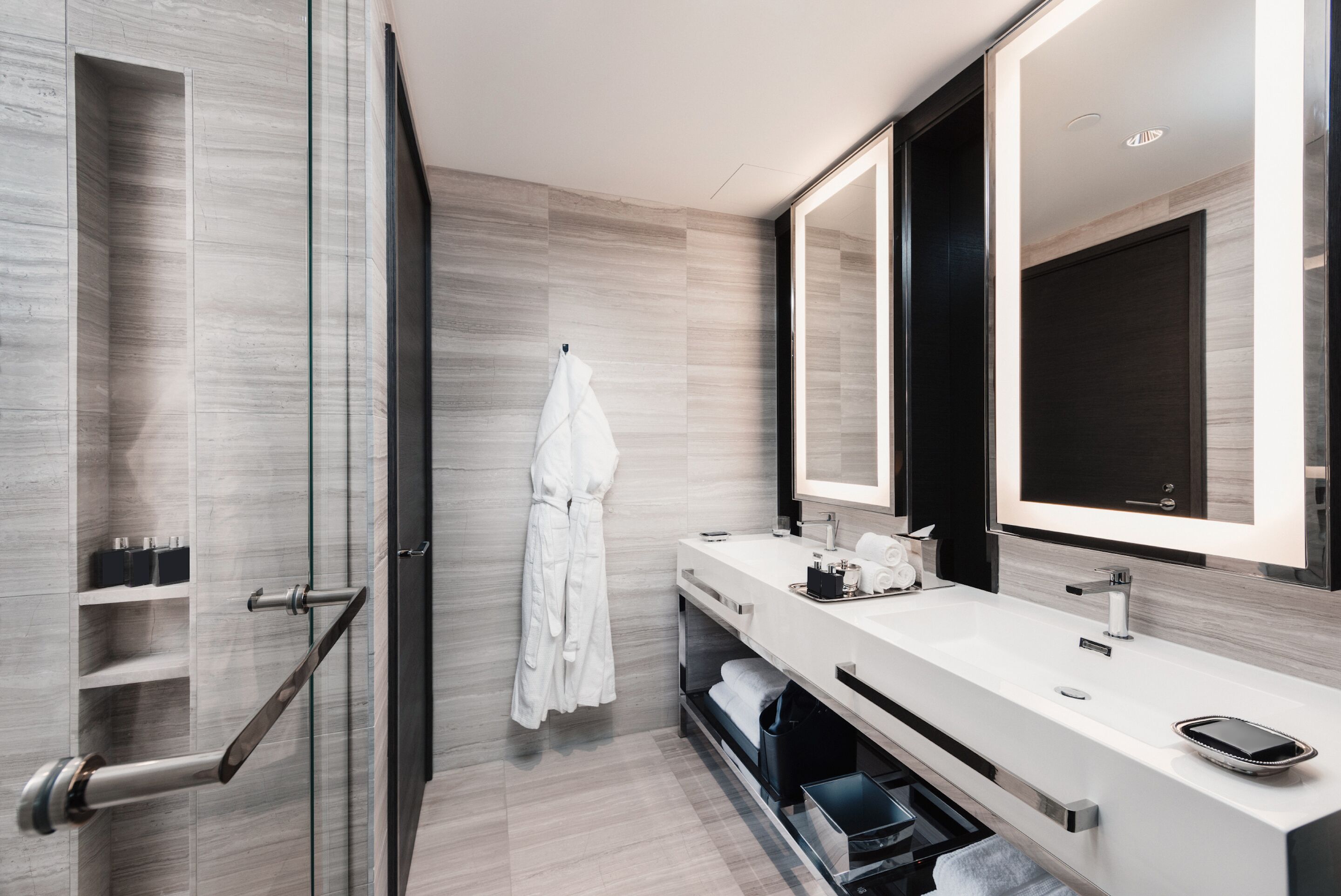 A modern bathroom with a glass shower enclosure, a white vanity with two sinks, a large mirror, and a towel rack with a white robe.