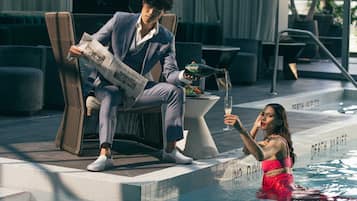 Un hombre leyendo un periódico junto a una piscina, una mujer con traje de baño rojo y una mesa con una botella y un vaso.