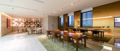 A modern library with a long wooden table and chairs, bookshelves, and a large window.