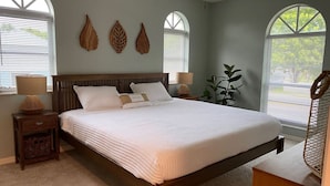 A bedroom with a large bed, two bedside tables, a nightstand, and a window with a view of greenery. - Sandy Feet Retreat - 3Bdrms, Pool, Steps to Beach (Fort Myers Beach)