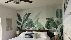 Une chambre avec un grand lit, un ventilateur de plafond et une fresque murale représentant de grandes feuilles.
