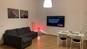 A modern living room with a black sofa, a dining table with four chairs, a flat-screen TV mounted on the wall, and a red lamp on a side table. - SUMMER WITH ME😁 (Bari)
