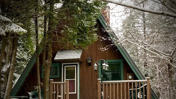 Una cabaña de madera con puerta y ventanas verdes, rodeada de árboles y cubierta de nieve.