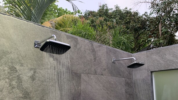 Une douche extérieure moderne, dotée d’un système de jets d’eau imitant la pluie, installée contre un mur à texture et entourée d’une végétation luxuriante.