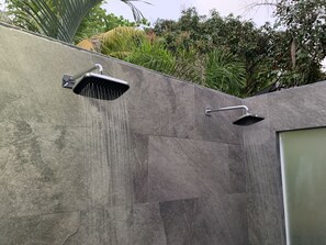 Eine moderne Außendusche mit regenduschartigem Wasserfall an einer strukturierten Wand, umgeben von üppigem Grün.