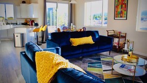 A modern living room with a blue sofa, yellow throw pillows, a glass coffee table, and a view of the kitchen and outside. - Kickin' It on Kindra! Modern 3 Bedroom New Build! (Cottonwood)