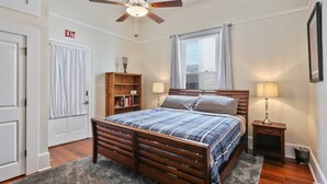 Une chambre à coucher avec un lit en bois, deux tables de chevet avec des lampes, un ventilateur de plafond, une fenêtre avec des rideaux et un panneau indiquant la sortie.