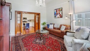 Un salon spacieux avec un tapis rouge à motifs, un canapé en cuir marron, un fauteuil gris et une table basse en verre. On y trouve un lustre, un tableau accroché au mur et, par une porte ouverte, on aperçoit la cuisine.