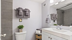 Ein modernes Badezimmer mit Toilette, Waschbecken und Dusche. Handtücher hängen an der Wand und ein gerahmtes Bild ist über der Toilette angebracht.