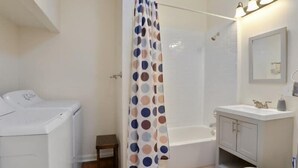 A bathroom with a white toilet, a bathtub, a shower curtain with a polka dot pattern, a small wooden stool, and a white washing machine. - Fab Studio | Walk to Food and Music | Pet Friendly (New Orleans)