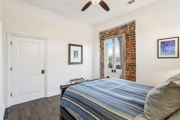 A bedroom with a bed, a door, a framed picture, a window with curtains, and a ceiling fan.