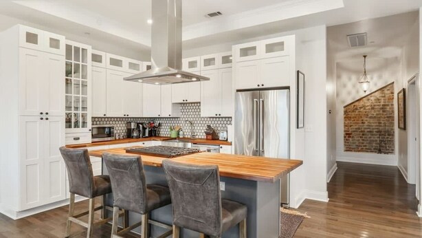 A modern kitchen with white cabinetry, a central island with bar stools, a stainless steel refrigerator, and a brick wall in the background.