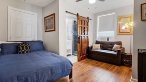 A bedroom with a large bed, a brown leather sofa, wooden flooring, a ceiling fan, and a sliding barn door.
