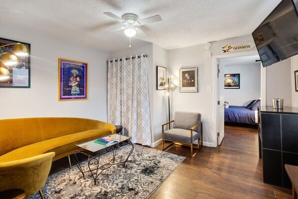 A living room with a yellow sofa, a glass coffee table, a patterned rug, a ceiling fan, and a wall-mounted television.