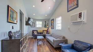 A spacious living area with a wooden floor, a sofa, a blue leather armchair, and a coffee table. There are framed pictures on the walls and an air conditioning unit mounted on the wall. - Charming Central 2BR | Courtyard | Pet Friendly (New Orleans)