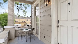 A covered porch with a table and chairs, a white door, and a view of a building in the background. - Lovely 3BD | Steps to St. Charles | Gated Parking (New Orleans)