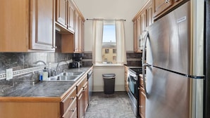 A compact kitchen with wooden cabinets, a stainless steel refrigerator, a sink, and a window with a view of buildings outside. - Lovely 3BD | Steps to St. Charles | Gated Parking (New Orleans)