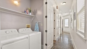 A laundry room with two front-loading washers and dryers, a shelf with towels and a potted plant, and a hallway with white doors and a tiled floor. - Lovely 3BD | Steps to St. Charles | Gated Parking (New Orleans)