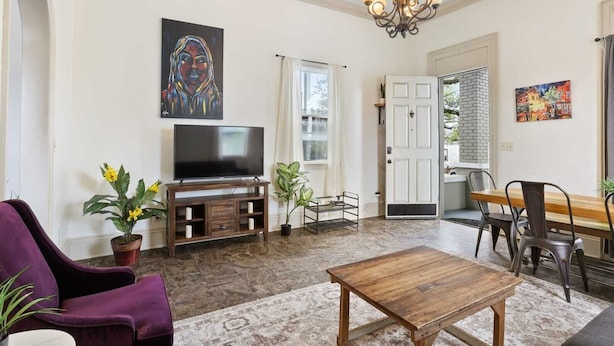 A living room with a TV, a wooden coffee table, purple armchairs, and a dining area with a wooden table and chairs. - Lovely 3BD | Steps to St. Charles | Gated Parking (New Orleans)