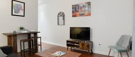 A modern living room with a wooden floor, a TV, a coffee table, a sofa, a chair, and a dining area with a table and chairs.