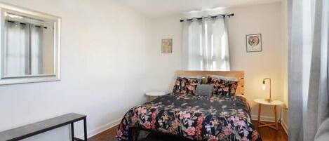 A bedroom with a bed, a bench, a ceiling fan, a window, and a small table with a lamp.