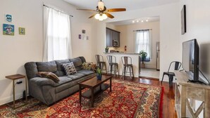 A living room with a sofa, coffee table, and a patterned rug. There is a ceiling fan, a TV, and a kitchen area in the background.