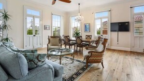 A spacious living room with a ceiling fan, a chandelier, a dining area with wooden chairs, and large windows.