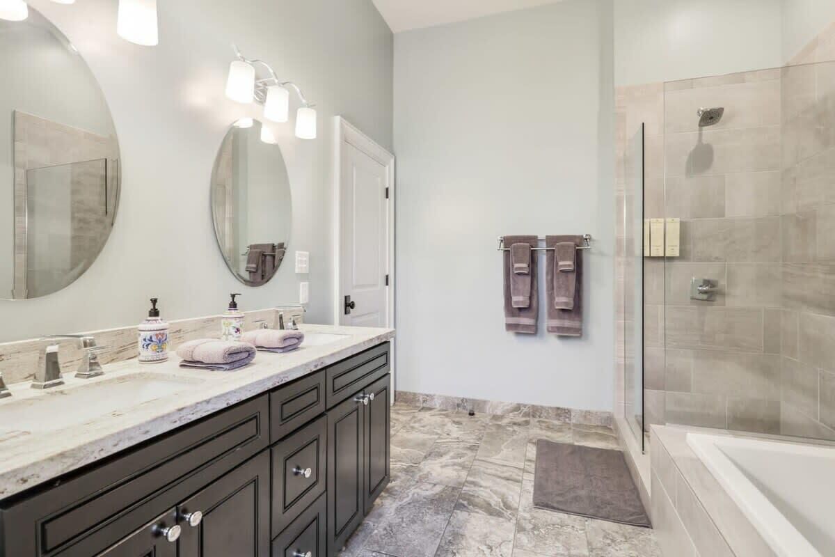 Une salle de bain moderne avec un grand miroir, deux vasques, des meubles sombres et une douche à l’italienne.