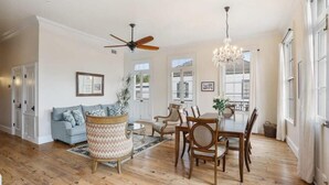 A spacious living room with hardwood flooring, a ceiling fan, a dining area with wooden chairs and a table, and a chandelier.