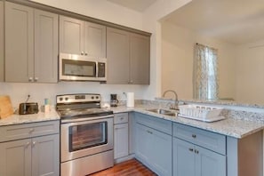 A kitchen with grey cabinets, stainless steel appliances, a granite countertop, and a window with patterned curtains.