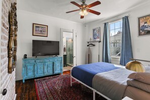 Une chambre à coucher avec un lit, une télévision, un ventilateur de plafond, une fenêtre avec des rideaux et un mur en briques.