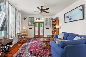 Ein Wohnzimmer mit einem blauen Sofa, einem Holztisch, einem Deckenventilator und einem roten Teppich mit Muster.