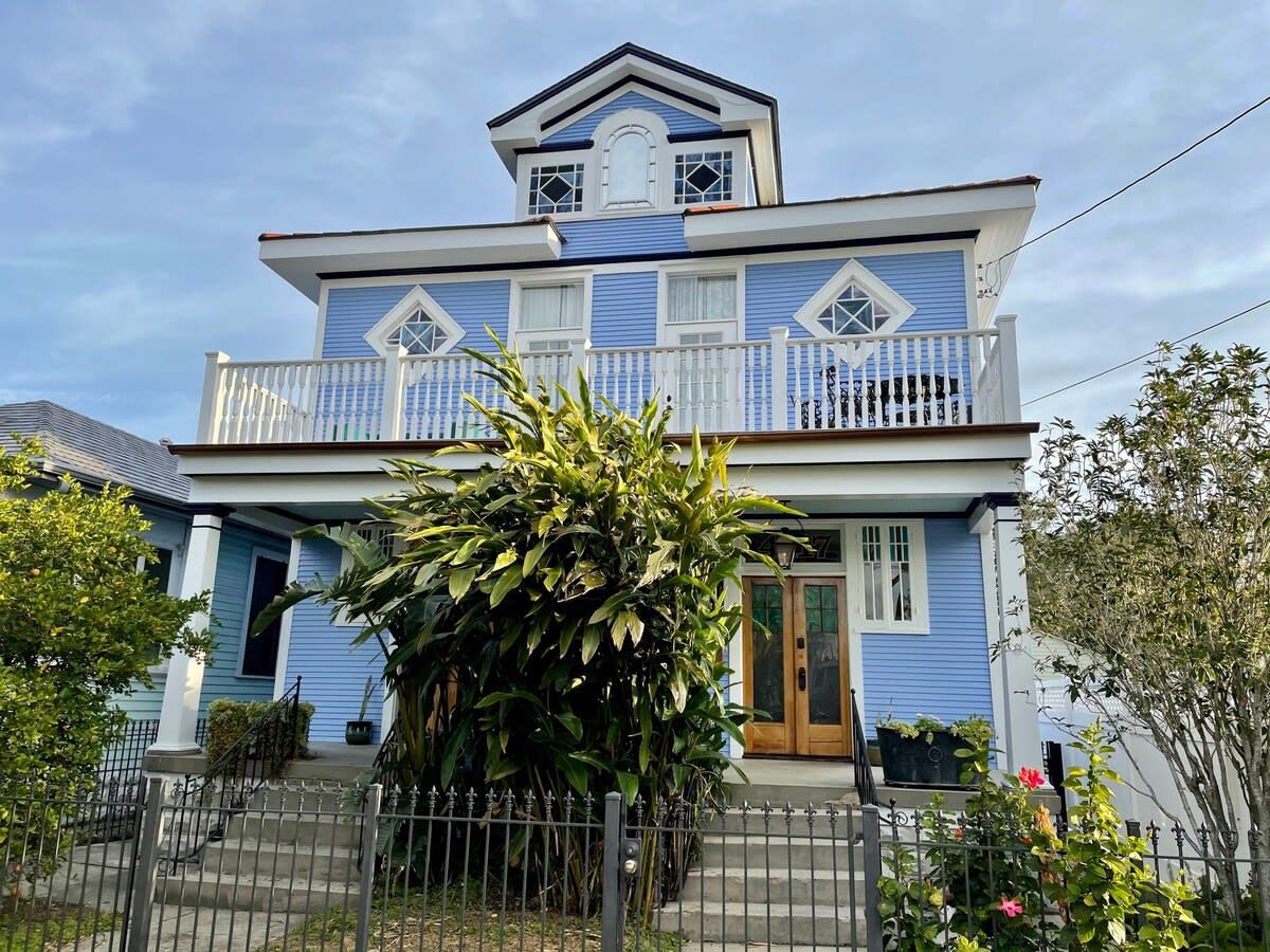 Une maison à deux étages, avec une façade bleue et des éléments décoratifs blancs, dotée d’un porche à l’avant et d’un petit jardin.