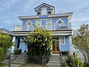Une maison à deux étages avec un balcon, une porte d’entrée et un grand arbre au premier plan.