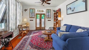 Un salon avec un canapé bleu, une table basse en bois, un tapis à motifs, un ventilateur de plafond et une bibliothèque.