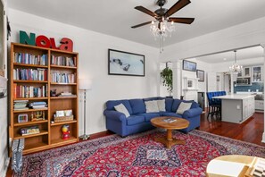 Un salon avec un canapé bleu, une bibliothèque en bois, un tapis à motifs et un ventilateur de plafond.