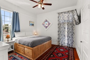 Een slaapkamer met een houten bed, een plafondventilator, een raam met blauwe gordijnen en een kamerbreed tapijt met een patroon.