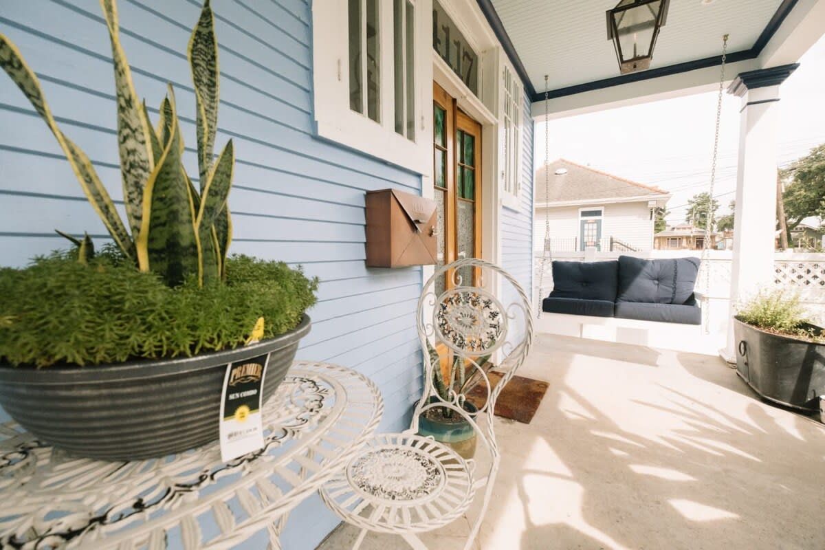 Eine überdachte Terrasse mit einem blauen Haus, einem weißen Metalltisch, einem Metallstuhl, einem hängenden Laternen und einem Briefkasten.